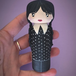 Puede incluir: Una mu&ntilde;eca de madera pintada a mano con cabello negro, una camisa blanca con cuello y un vestido negro con lunares blancos. La mu&ntilde;eca tiene una boca roja y ojos negros.