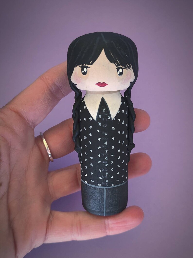 Puede incluir: Una mu&ntilde;eca de madera pintada a mano con cabello negro, una camisa blanca con cuello y un vestido negro con lunares blancos. La mu&ntilde;eca tiene una boca roja y ojos negros.