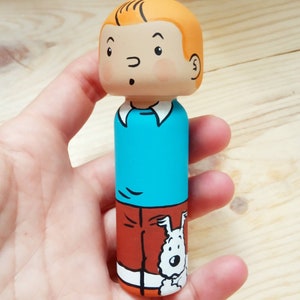 May include: A hand-painted wooden doll of a cartoon character with orange hair, a blue shirt, orange pants, and a white dog peeking out from behind the pants.