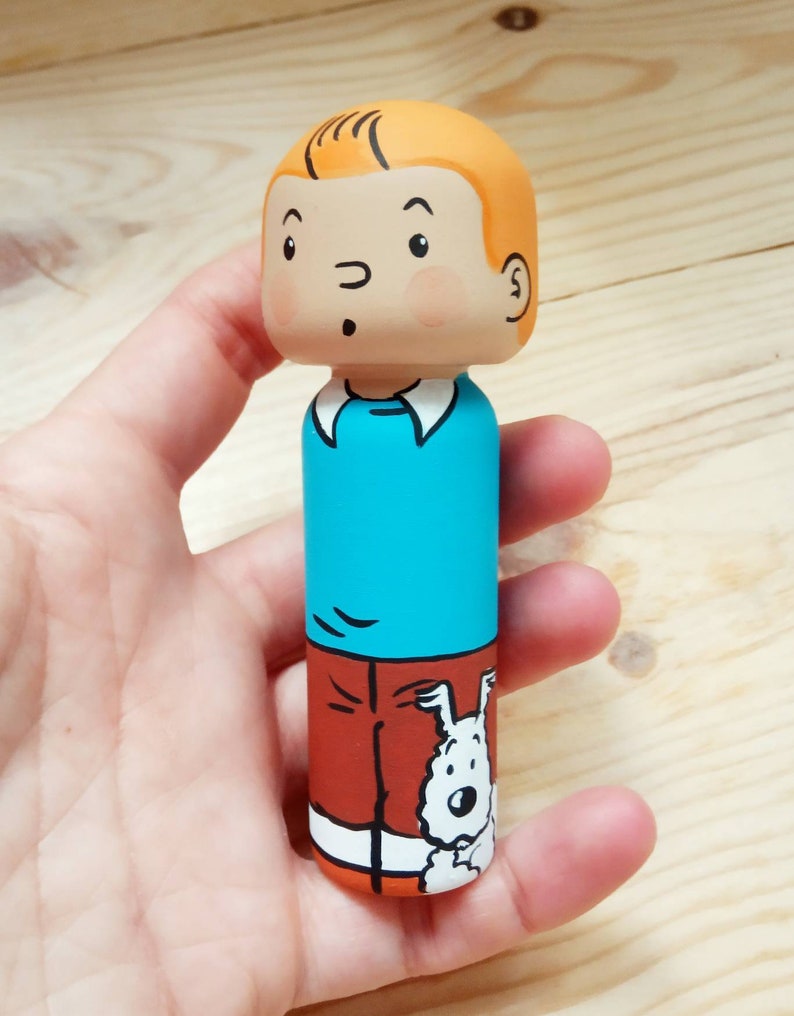 May include: A hand-painted wooden doll of a cartoon character with orange hair, a blue shirt, orange pants, and a white dog peeking out from behind the pants.