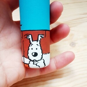 May include: A hand holds a small, cylindrical container with a blue, orange, and white cartoon illustration of a dog. The dog is white with black eyes and a black nose. The dog is wearing a red collar.