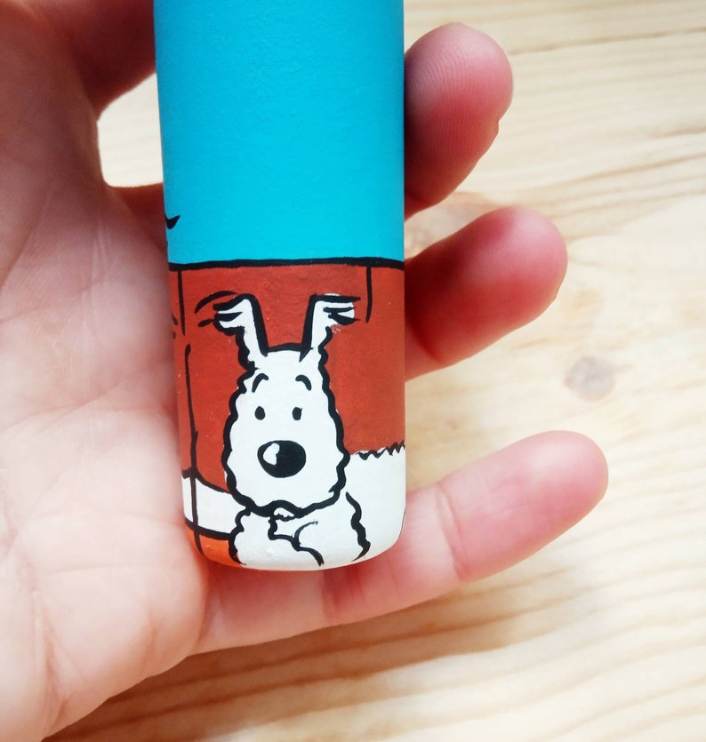 May include: A hand holds a small, cylindrical container with a blue, orange, and white cartoon illustration of a dog. The dog is white with black eyes and a black nose. The dog is wearing a red collar.