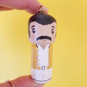Puede incluir: Un llavero de madera con una caricatura pintada a mano de un hombre con bigote, que lleva una chaqueta amarilla y pantalones blancos. El llavero tiene un anillo de metal dorado.