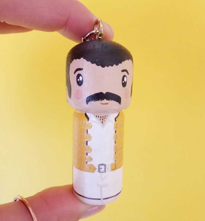 Puede incluir: Un llavero de madera con una caricatura pintada a mano de un hombre con bigote, que lleva una chaqueta amarilla y pantalones blancos. El llavero tiene un anillo de metal dorado.