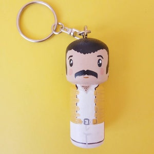 Puede incluir: Un llavero de madera con una caricatura pintada a mano de un hombre con bigote, que lleva una chaqueta amarilla y pantalones blancos. El llavero tiene un anillo de metal plateado.