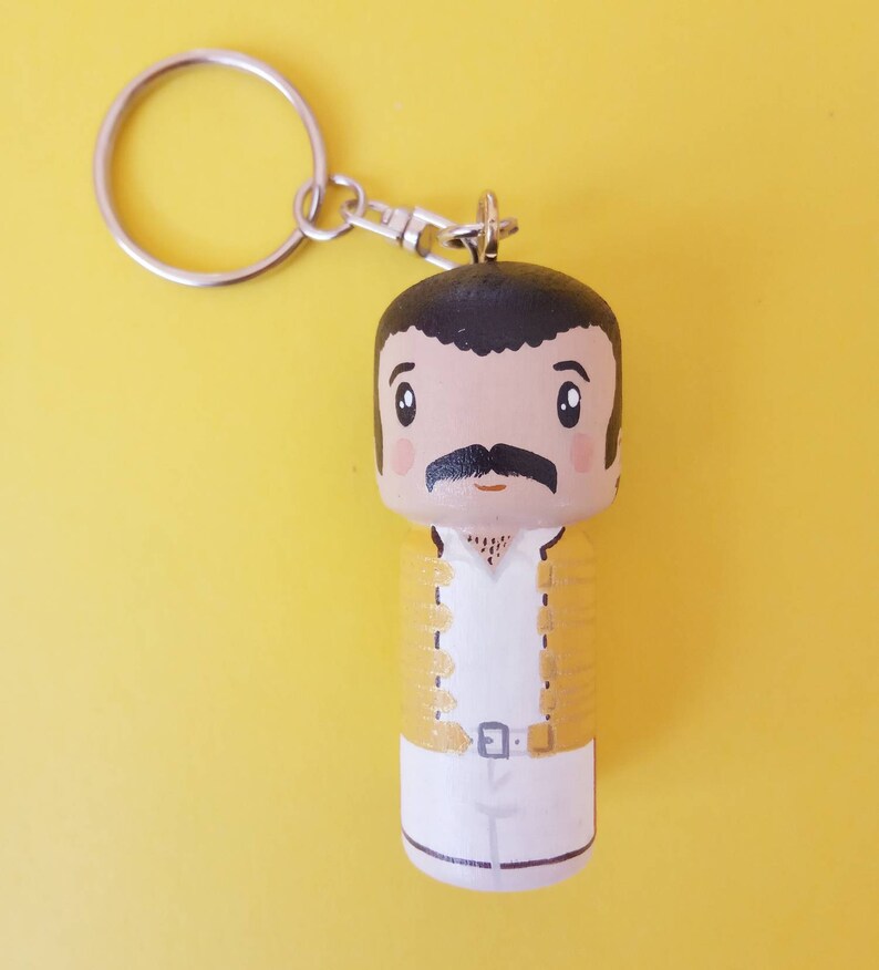 Puede incluir: Un llavero de madera con una caricatura pintada a mano de un hombre con bigote, que lleva una chaqueta amarilla y pantalones blancos. El llavero tiene un anillo de metal plateado.
