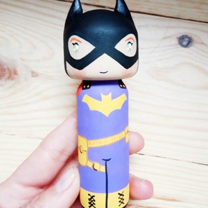 K&ouml;nnte beinhalten: Eine Holzpuppe, die wie Batgirl bemalt ist. Die Puppe hat eine schwarze Maske, einen lila Anzug, einen gelben G&uuml;rtel und gelbe Stiefel. Die Puppe h&auml;lt ein Fledermaussymbol.