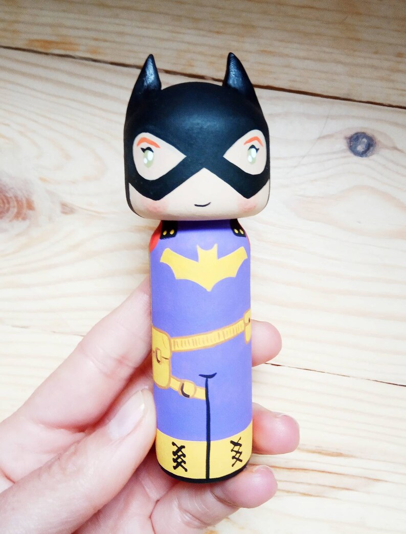 K&ouml;nnte beinhalten: Eine Holzpuppe, die wie Batgirl bemalt ist. Die Puppe hat eine schwarze Maske, einen lila Anzug, einen gelben G&uuml;rtel und gelbe Stiefel. Die Puppe h&auml;lt ein Fledermaussymbol.