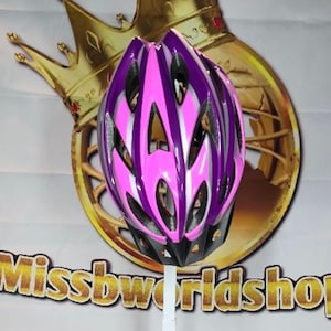 May include: A pink and purple bicycle helmet with a black base and silver accents. The helmet has multiple vents for ventilation and is designed for safety. The background includes a gold crown and the text "Missbworldshop".