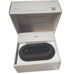May include: A dark gray oval-shaped speaker with control buttons, nestled inside a white box. The box is open, revealing the speaker and its packaging. The top of the box has the Microsoft logo and text indicating compatibility with Windows 10/11 and macOS.