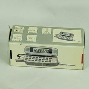 Puede incluir: Una combinación de teléfono y calculadora vintage en su embalaje original. La caja es de color beige claro con una parte superior gris y presenta ilustraciones de material de oficina. El teléfono-calculadora es gris con un teclado negro y una pantalla digital que muestra los números 12345678.
