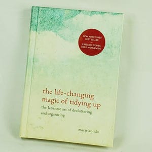 Puede incluir: Un libro titulado "the life-changing magic of tidying up" de Marie Kondo. La portada presenta un diseño de acuarela en tonos verdes y blancos. Una pegatina roja indica que es un best-seller del New York Times.