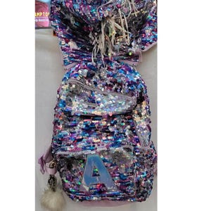 May include: A sequined backpack in shades of purple, blue, and silver. The backpack features a large letter "A" on the front and a fluffy white pom-pom charm. The bag has a top handle and adjustable straps.