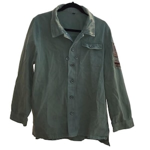 May include: A long-sleeved, button-down shirt in a muted green hue. The shirt features a classic collar, a chest pocket, and decorative stitching on the right shoulder. The shirt is hanging on a black hanger.