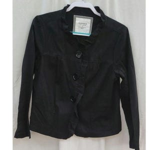 May include: Black Sonoma jacket with a frilled collar and three large buttons. The jacket has long sleeves and two front pockets. A tag is visible on the collar.