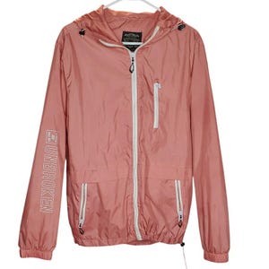 May include: A dusty rose windbreaker jacket with a hood, white zippers, and two front pockets. The jacket has the word "UNBROKEN" printed vertically on the left sleeve. The jacket is hanging on a white hanger.