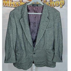Könnte beinhalten: Ein grauer Tweed-Blazer mit Zwei-Knopf-Verschluss, fallendem Revers und zwei Vordertaschen. Der Blazer hat ein lila Futter und hängt an einem weißen Kleiderbügel. Klassischer Stil.