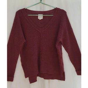 May include: A burgundy V-neck sweater with long sleeves. The sweater has a textured knit design and a small tag near the neckline. The sleeves are cuffed at the wrists. The sweater is hanging on a green hanger against a white background.
