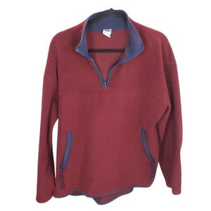 May include: A burgundy fleece pullover with a navy blue collar, zipper, and pocket trim. The long-sleeved top has a half-zip closure and two front pockets. The garment is hanging on a black hanger.