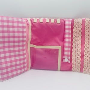 Op de afbeelding: Een roze en wit geruite stoffen organizer met een vak met ritssluiting, een doorzichtig plastic zakje, een reeks kleine stoffen lussen, een bandje met drukknoop en twee verticale stroken decoratief crèmekleurig kant.