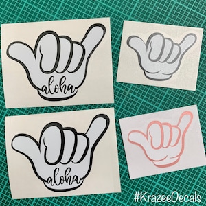 May include: Four white vinyl decals with a hand gesture design. Two decals feature the word "aloha" in a script font. One decal is a light gray outline, and another is a rose gold color. The decals are on a green cutting mat.