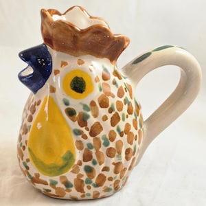 May include: A ceramic pitcher shaped like a rooster with a glossy finish. The body features brown and green spots on a cream background, a bright yellow wattle, a dark blue beak, and a brown comb. It includes a curved handle on the side.