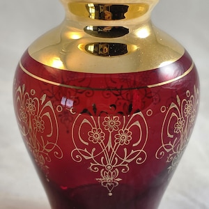 May include: A ruby red glass vase featuring a wide gold-colored neck and rim. The body of the vase is decorated with intricate gold-colored floral patterns and scrolling lines. The base of the vase is clear glass with a gold-colored band accent.