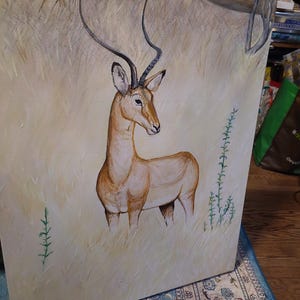 Op de afbeelding: Een olieverfschilderij op doek met een impala die in hoog, lichtgekleurd gras staat. De impala heeft een beige vacht, donkere gebogen hoorns en staat onder een grote, overhangende boomtak. De achtergrond heeft gedempte gele tinten.