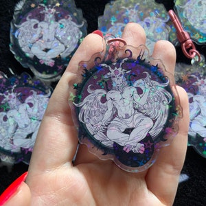 May include: A holographic charm featuring a white, winged, goat-like figure with horns and a halo. The figure is surrounded by stars and a black background. The charm is made of clear plastic and has a shiny, iridescent finish.