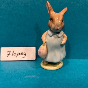 Puede incluir: Figura de cerámica de un conejo con un vestido azul claro y una bolsa rosa. El conejo tiene pelaje marrón y está de pie sobre una pequeña base redonda amarilla. El texto "7lopsy" está en un rectángulo blanco.