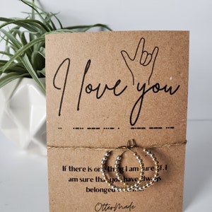 May include: A brown card with the text "I love you" in black script and a hand sign for "I love you" in black. The card has a string tied around it with two gold and silver hoop earrings hanging from the string. The card also has the text "If there is one thing I am sure of, I am sure that you have always belonged here." and "Otter Made" in black script.