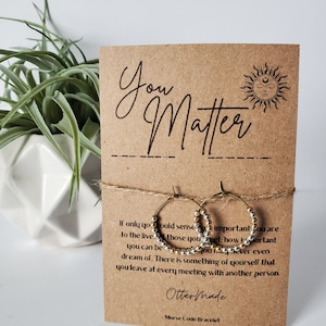 May include: A pair of gold and silver hoop earrings with a brown card that reads "You Matter" and "Otter Made Morse Code Bracelet".
