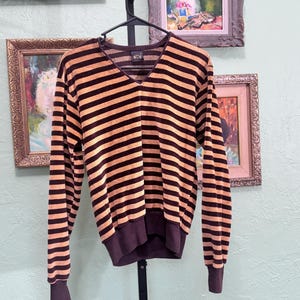 May include: A long-sleeved, V-neck sweater with horizontal stripes in brown and tan. The sweater is hanging on a black hanger against a light green wall. The cuffs and hem are a darker brown.