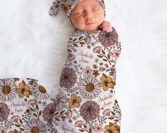 May include: A white swaddle with a floral pattern in shades of pink, brown, and yellow. The swaddle has the name "Anabelle June" printed on it. A matching floral hat is also included.