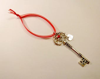 May include: A silver key with an ornate design and a red heart charm. The key is attached to a red ribbon.