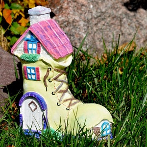 May include: A whimsical yellow boot with a pink and white house on top. The house has a chimney and windows. The boot is sitting in green grass.