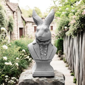 Op de afbeelding: Grijze betonnen buste van een konijn met een jasje en een gekraagde kraag. De konijnensculptuur heeft lange oren en is gemonteerd op een stenen voet. De buste staat buiten in een tuin.