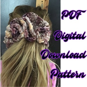 May include: A digital download pattern for a crocheted hair scrunchie. The scrunchie is made with a velvet yarn in shades of purple, pink, and beige. The text "PDF Digital Download Pattern" is written in black with a purple outline.