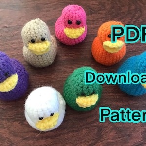 May include: A set of seven crocheted duck plushies in different colors, including purple, gray, pink, orange, green, white, and blue. The ducks have yellow beaks and black eyes. The text "PDF Download Pattern" is superimposed on the image.