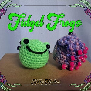 May include: Two crocheted frog toys. One is green with black eyes and a smile. The other is purple, pink, and white with black eyes and pink beads around the edges. The text "Fidget Frogs" is written in green above the toys. The text "Art by Heather" is written below the toys.