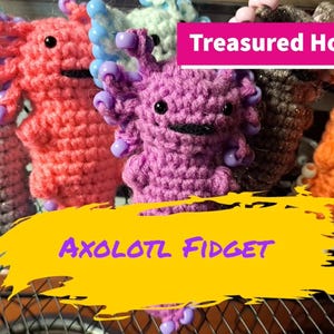 May include: Five crocheted axolotl plushies with colourful beads. The axolotls are in various colours, including grey, orange, purple, and white. The text "Treasured Hometown" and "Axolotl Fidget" is visible in the image.