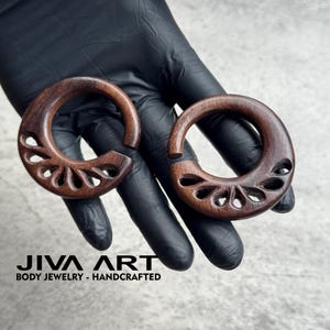 May include: Two dark brown wooden ear gauges with a carved floral design. The gauges are crescent-shaped and held in a gloved hand. The text "JIVA ART BODY JEWELRY - HANDCRAFTED" is at the bottom.