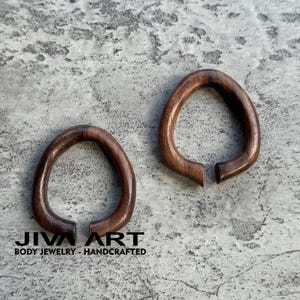 May include: Two dark brown, teardrop-shaped wooden body jewelry pieces. The jewelry is open at the bottom and has a smooth, polished finish. The text "JIVA ART BODY JEWELRY - HANDCRAFTED" is at the bottom.