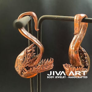 May include: A pair of copper-coloured body jewellery earrings. Each earring features a curved design with a detailed, open-mouthed creature head. The earrings are displayed on a black metal stand. The text "JIVA ART BODY JEWELRY - HANDCRAFTED" is visible.