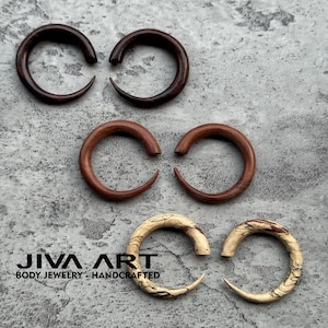 May include: Three pairs of wooden ear gauges in varying shades of brown and tan. The gauges are crescent-shaped, with a smooth, polished finish. The bottom pair has a unique, patterned grain. The text "JIVA ART BODY JEWELRY - HANDCRAFTED" is at the bottom.