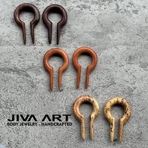 May include: A selection of handcrafted wooden body jewellery, including ear gauges and plugs. The pieces are in various shades of brown, from dark to light, and feature a unique, curved design. The brand name "JIVA ART" is visible.