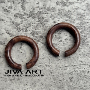 May include: Two dark brown wooden ear gauges with a circular design and a small opening. The gauges are handcrafted and have a smooth finish. The text "JIVA ART BODY JEWELRY - HANDCRAFTED" is visible at the bottom.