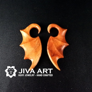 May include: A pair of wooden ear gauges shaped like wings. The gauges are a light brown color and have a smooth finish. JIVA ART BODY JEWELRY - HAND CRAFTED is printed below the gauges.