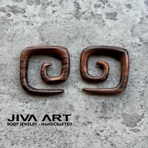 May include: Pair of dark brown wooden spiral-shaped earrings. The earrings are square with a rounded spiral design. The text "JIVA ART BODY JEWELRY - HANDCRAFTED" is printed in black at the bottom of the image.