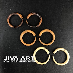 May include: Three pairs of wooden ear gauges in different wood grain patterns. The top pair is dark brown with black streaks, the middle pair is light brown, and the bottom pair is light brown with dark brown streaks. JIVA ART BODY JEWELRY - HANDCRAFTED is printed below the gauges.